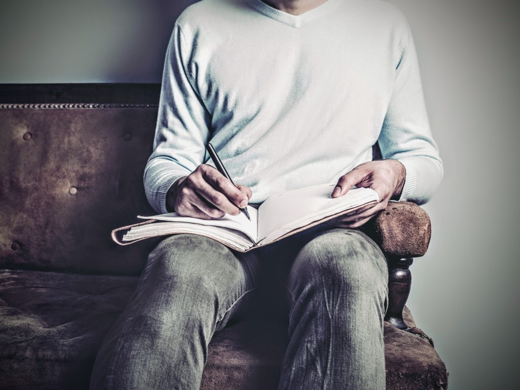 A young man is sitting on an old sofa and is writing in a notebook